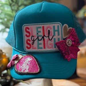 May include: Teal trucker hat with a mesh back and a curved brim. The hat features a patch with the words "SLEIGH girl SLEIGH" in red, pink, and teal. Embellishments include a pink sequined bow with a rhinestone center and a pink sequined strawberry.