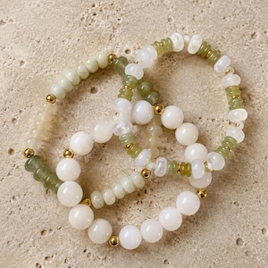May include: Two elastic bracelets featuring a mix of smooth white round beads, translucent pale green disc beads, and small polished gold-tone metal spacer beads. The bracelets are arranged in an overlapping circular pattern on a textured surface.