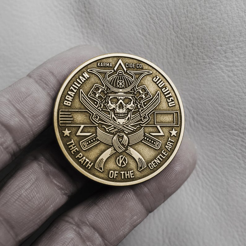 May include: A gold-colored coin with a detailed design featuring a skull wearing a samurai helmet, crossed swords, and the words "Brazilian Jiu-Jitsu" and "The Path of the Gentle Art". The coin also has the text "Karma Cide Co".