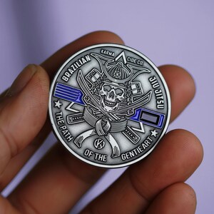 May include: A silver-colored coin with a skull wearing a samurai helmet, crossed swords, and the words "Brazilian Jiu Jitsu." The coin also features the text "Karma" and "Cide Co.", with a blue stripe design.