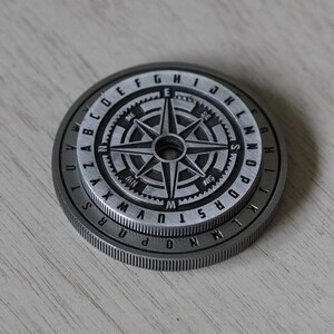 Peut inclure: Une pi&egrave;ce de monnaie en m&eacute;tal argent&eacute; avec un motif de boussole et un anneau de lettres autour du bord. La boussole pr&eacute;sente une &eacute;toile noire &agrave; quatre branches et les lettres N, S, E et W.