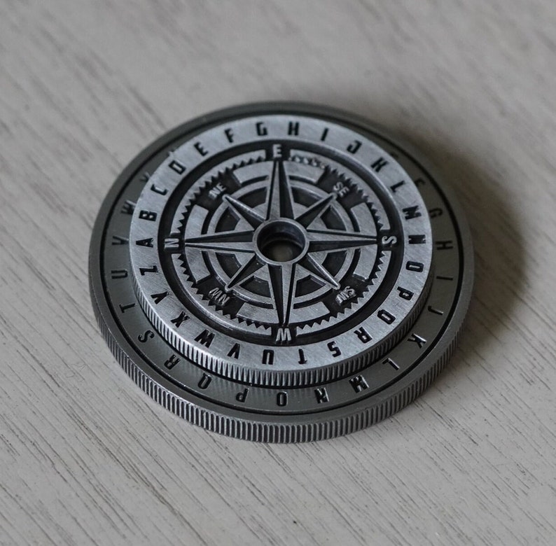 Peut inclure: Une pi&egrave;ce de monnaie en m&eacute;tal argent&eacute; avec un motif de boussole et un anneau de lettres autour du bord. La boussole pr&eacute;sente une &eacute;toile noire &agrave; quatre branches et les lettres N, S, E et W.