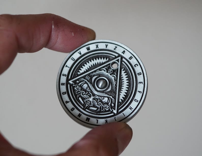 Peut inclure: Un disque en m&eacute;tal argent&eacute; avec un motif circulaire repr&eacute;sentant un &oelig;il stylis&eacute; &agrave; l'int&eacute;rieur d'un triangle. Le disque est entour&eacute; d'un anneau de lettres, commen&ccedil;ant par "Q" et allant dans le sens des aiguilles d'une montre jusqu'&agrave; "Z".