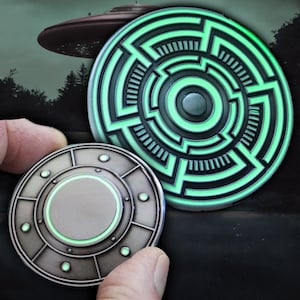 May include: Two glow-in-the-dark metal UFO-shaped coins. One coin features a circular maze design with a green glow. The other coin has a smooth, circular center with a green glow.