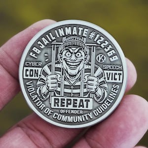 May include: A silver-colored coin with a cartoon illustration of a grinning inmate behind bars. The coin has the text "FB JAIL INMATE #122369", "CYBER CON", "SPEECH VICT", "VIOLATOR OF COMMUNITY GUIDELINES", and "REPEAT OFFENDER".