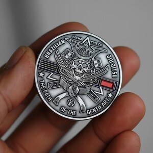 May include: A silver-colored coin with a skull wearing a samurai helmet and crossed swords. The coin features the words "Brazilian Jiu Jitsu", "The Path of the Gentle Art", and "Karma Cide Co."