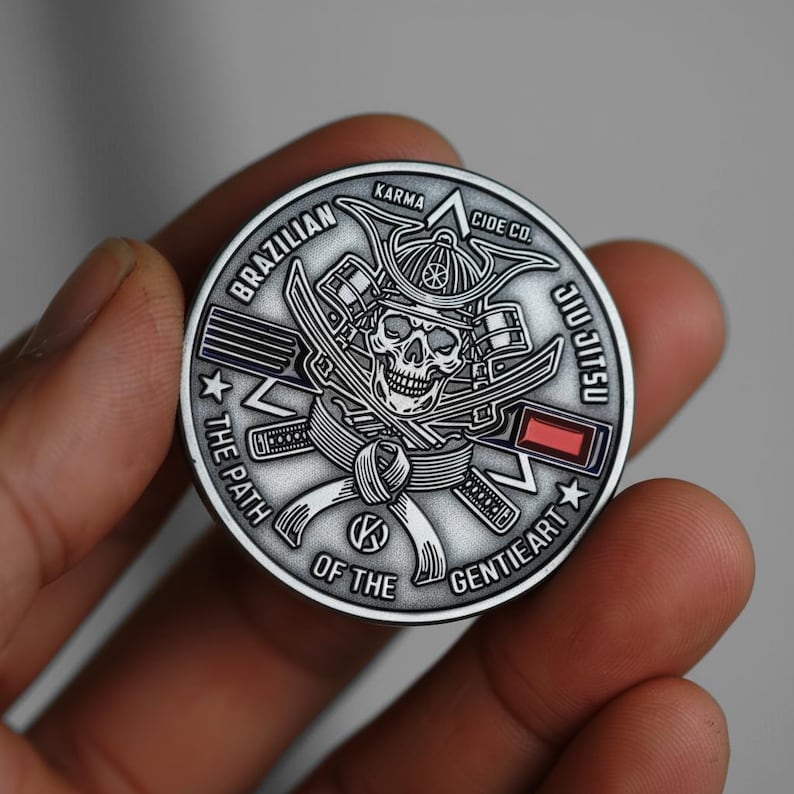May include: A silver-colored coin with a skull wearing a samurai helmet and crossed swords. The coin features the words "Brazilian Jiu Jitsu", "The Path of the Gentle Art", and "Karma Cide Co."