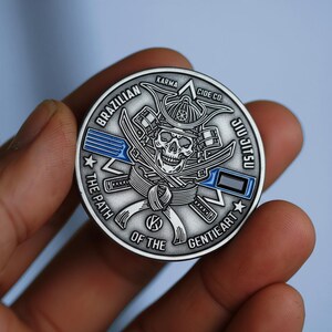 May include: A silver coin featuring a skull wearing a samurai helmet, crossed swords, and the text "BRAZILIAN JIU JITSU." The coin also includes the words "KARMA," "CIDE CO," and "THE PATH OF THE GENTIE ART."
