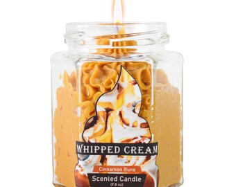 May include: A lit, scented candle in a clear, square jar. The candle is a light brown color, with a label that reads "Whipped Cream Cinnamon Buns Scented Candle (9.8 oz)". The candle has a decorative design and a lit wick.