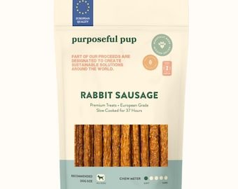 May include: A packet of Purposeful Pup Rabbit Sausage dog treats. The bag is white with green and blue details, and features text including "Rabbit Sausage" and "Premium Treats." The treats are brown, sausage-shaped, and visible through the clear section.