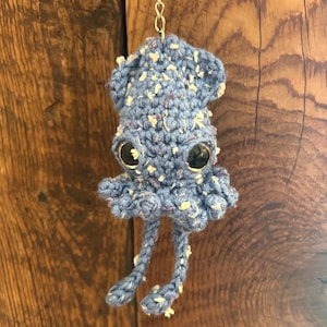 May include: A handmade, blue crochet squid with large, black, googly eyes. The squid has a textured appearance with white and purple flecks throughout its body. It is suspended by a silver chain, showcasing its whimsical design.