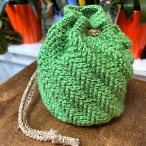 May include: A green crocheted drawstring pouch with a textured weave pattern. The pouch has a beige drawstring closure. The pouch is sitting on a wooden surface. The background includes potted plants.