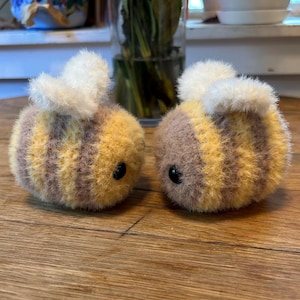 May include: Two plush bee toys with yellow and brown striped bodies and fluffy white wings. Each bee has a pair of small, black, round eyes. The bees are sitting on a wooden surface.