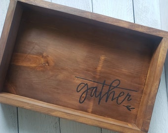 May include: A rectangular wooden tray with a dark brown stain. The word "gather" is written in a cursive font in black ink across the tray's surface. A small leaf design is next to the word.