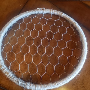 May include: A circular craft hoop featuring a chicken wire mesh center. The outer ring is wrapped with natural-colored twine. The metal wire is silver, and the twine is a light beige. This item is suitable for DIY projects and home decor.