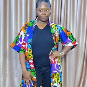 May include: A colorful, patterned kimono-style jacket with short sleeves. The jacket features a vibrant mix of colors including blue, green, orange, and purple. The jacket is worn over a black top and black pants. The brand name "NIKDIVA FASHION" is visible.