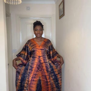 May include: A long, flowing dress with a V-neck and wide sleeves. The dress has a vibrant tie-dye pattern in shades of orange and navy blue. The fabric appears lightweight, suitable for warmer weather.