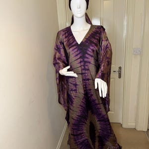 May include: A long, flowing dress and matching head covering in shades of purple and brown, displayed on a mannequin. The fabric features a tie-dye pattern. The dress has a V-neck and wide sleeves. The head covering is a close-fitting cap.