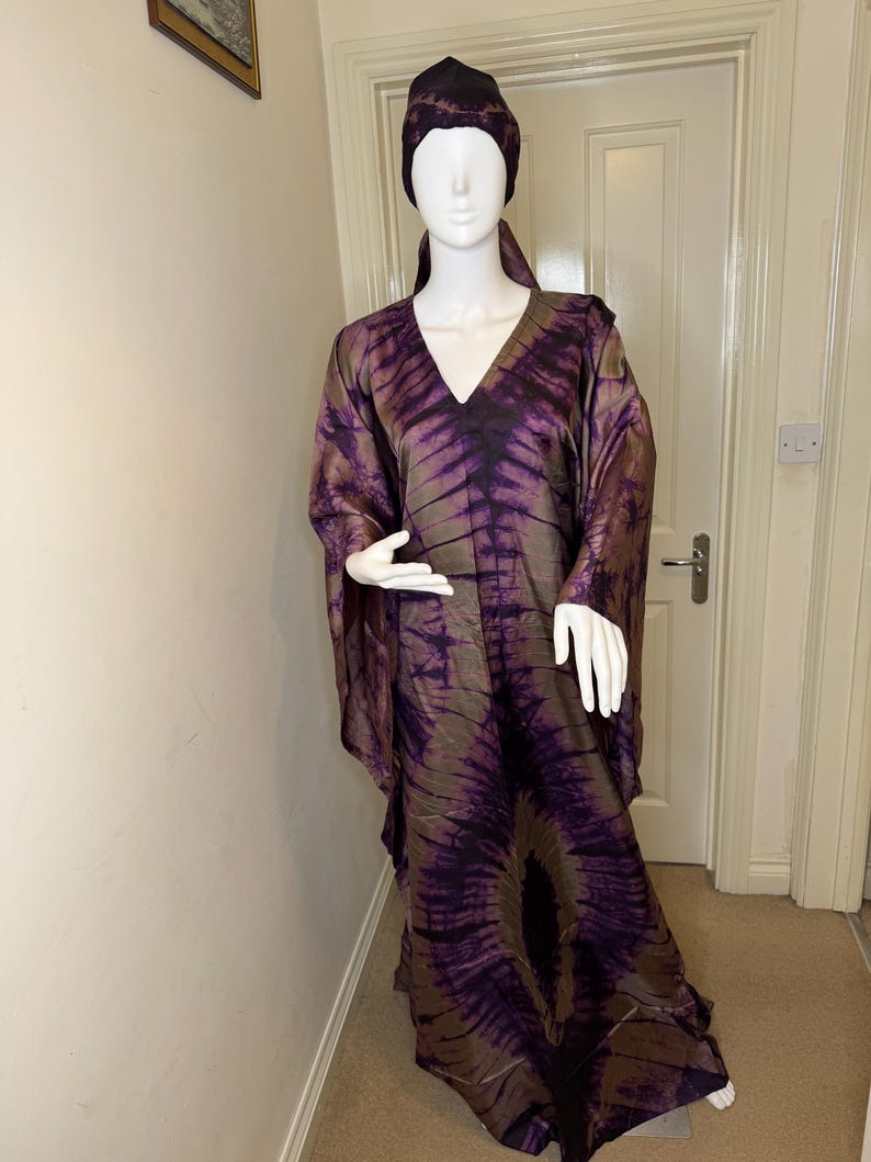 May include: A long, flowing dress and matching head covering in shades of purple and brown, displayed on a mannequin. The fabric features a tie-dye pattern. The dress has a V-neck and wide sleeves. The head covering is a close-fitting cap.