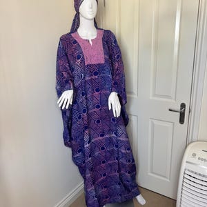 May include: A long, flowing dress and matching head covering in blue and purple with a geometric pattern are displayed on a mannequin. The dress has a pink embroidered neckline. The mannequin stands in front of a white door. The fabric has a geometric design.