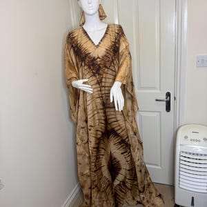 May include: A flowing, tie-dye patterned dress and matching head wrap in shades of brown and tan. The dress features a V-neck and wide sleeves. The fabric has a soft, flowing appearance, creating a relaxed and elegant look. The outfit is displayed on a mannequin.