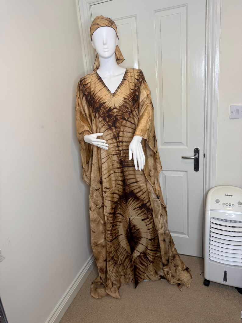May include: A flowing, tie-dye patterned dress and matching head wrap in shades of brown and tan. The dress features a V-neck and wide sleeves. The fabric has a soft, flowing appearance, creating a relaxed and elegant look. The outfit is displayed on a mannequin.