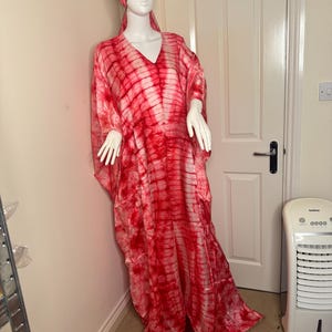 May include: A red and white tie-dye caftan and matching head wrap displayed on a mannequin. The flowing, V-neck caftan is floor-length and features a bold, striped pattern. The head wrap mirrors the caftan's design, creating a cohesive ensemble. The outfit is displayed indoors.