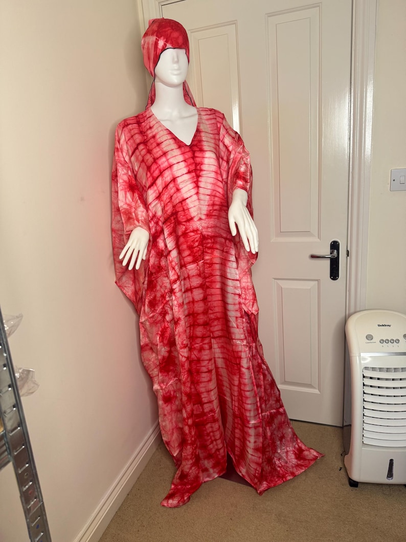 May include: A red and white tie-dye caftan and matching head wrap displayed on a mannequin. The flowing, V-neck caftan is floor-length and features a bold, striped pattern. The head wrap mirrors the caftan's design, creating a cohesive ensemble. The outfit is displayed indoors.