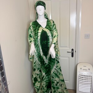 May include: A green and white tie-dye caftan dress and matching head covering on a mannequin. The dress has a V-neck and wide sleeves. The fabric has a pattern of dark green and white swirls.