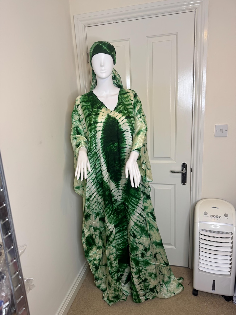 May include: A green and white tie-dye caftan dress and matching head covering on a mannequin. The dress has a V-neck and wide sleeves. The fabric has a pattern of dark green and white swirls.