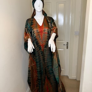 May include: A tie-dye caftan and matching head wrap in shades of green, brown, and orange are displayed on a mannequin. The caftan has a V-neck and flowing sleeves. The fabric has a marbled effect.