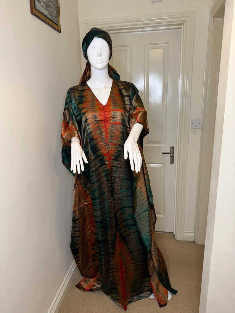 May include: A tie-dye caftan and matching head wrap in shades of green, brown, and orange are displayed on a mannequin. The caftan has a V-neck and flowing sleeves. The fabric has a marbled effect.