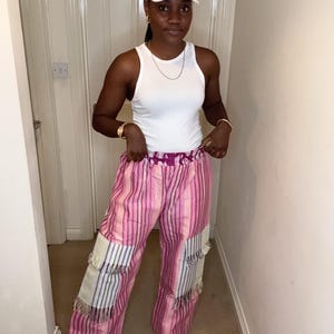 May include: A white vest top and pink striped trousers with fringed patches. The outfit is completed with a white baseball cap with "YU" on it and white trainers. A gold necklace and watch are also visible. The outfit is worn indoors.