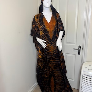 May include: A long, flowing black and orange patterned caftan dress with a matching head wrap. The dress features a V-neck and wide sleeves. The fabric has a textured appearance.