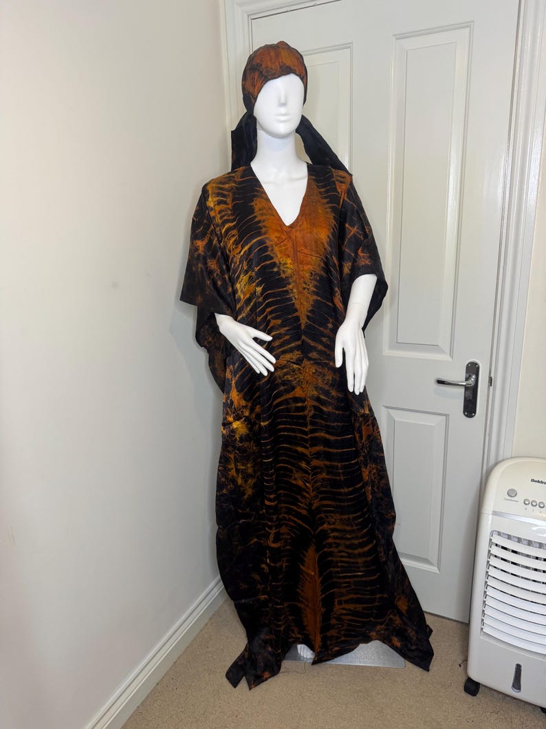 May include: A long, flowing black and orange patterned caftan dress with a matching head wrap. The dress features a V-neck and wide sleeves. The fabric has a textured appearance.