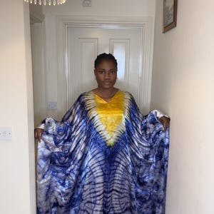 May include: A flowing, tie-dye caftan dress in shades of blue and white, with a yellow accent at the neckline. The dress has a V-neck and wide sleeves. The fabric appears soft and lightweight, suitable for warm weather.