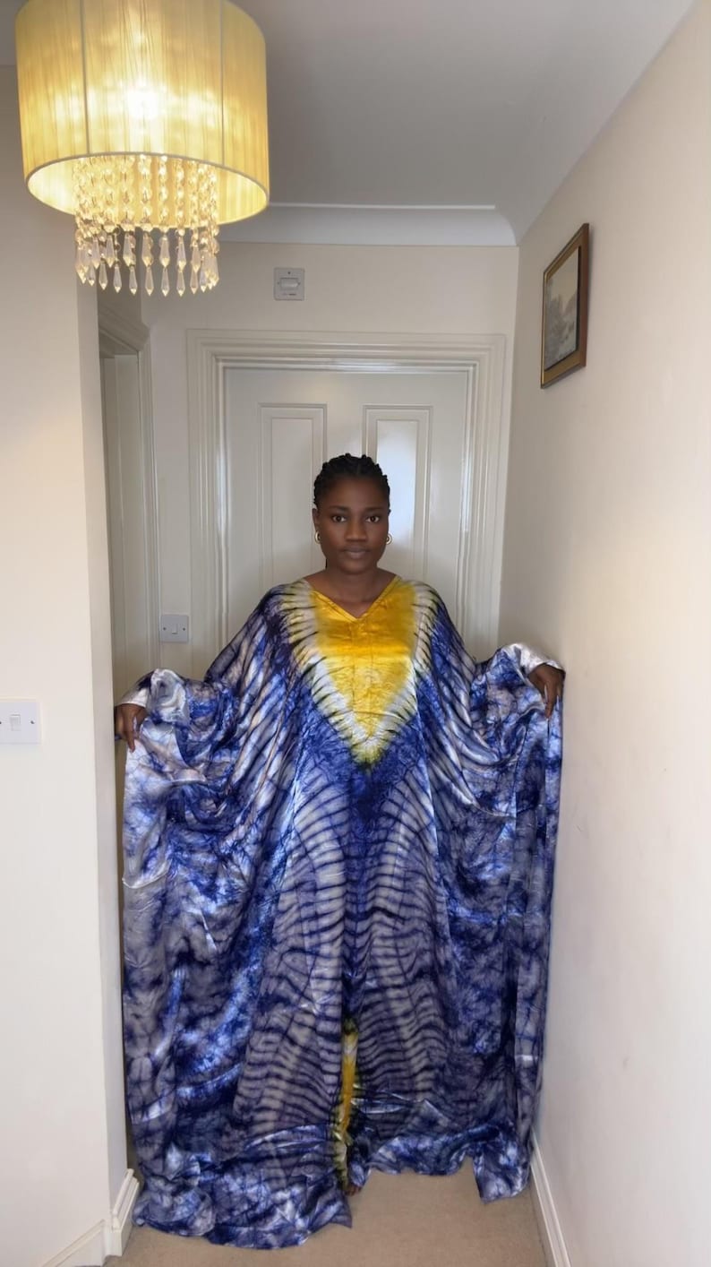 May include: A flowing, tie-dye caftan dress in shades of blue and white, with a yellow accent at the neckline. The dress has a V-neck and wide sleeves. The fabric appears soft and lightweight, suitable for warm weather.