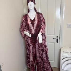 May include: A burgundy and white tie-dye caftan and matching head wrap displayed on a mannequin. The caftan has a V-neck and flowing sleeves. The fabric has a vertical stripe pattern.