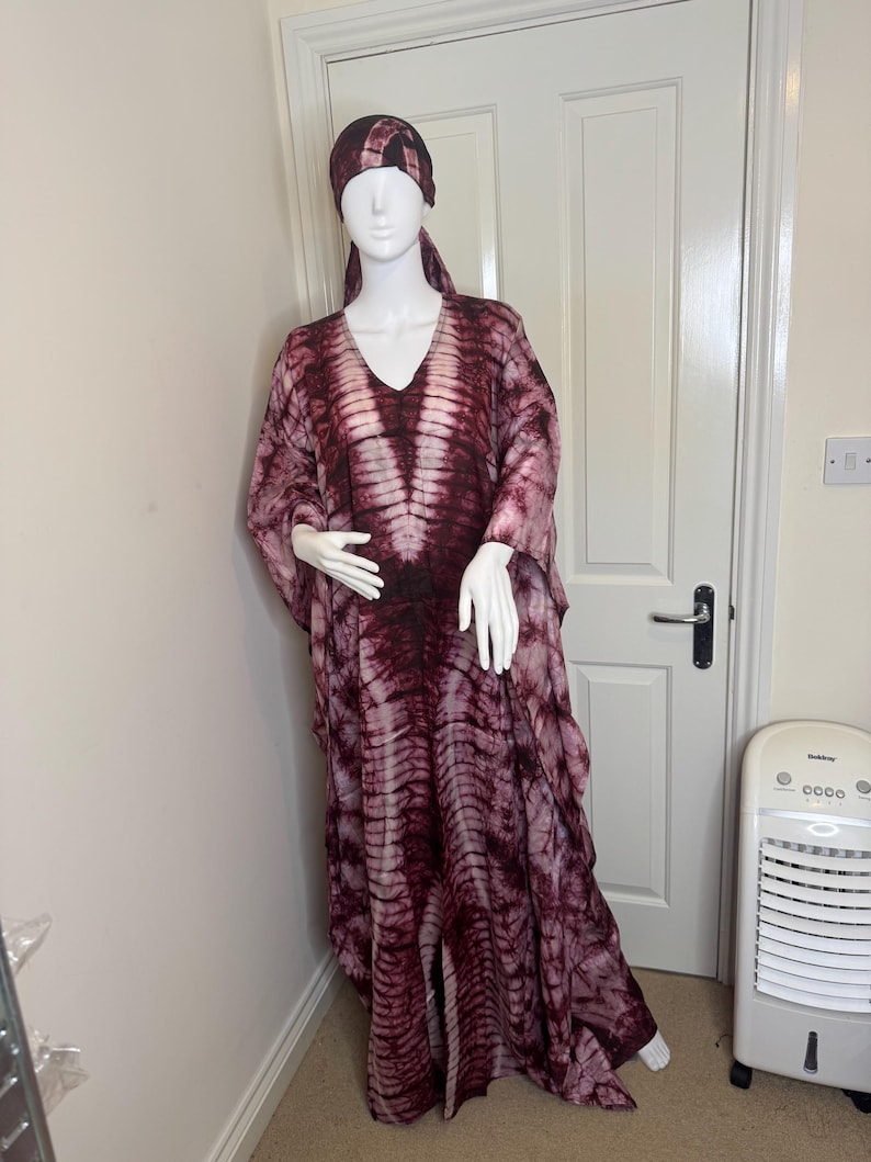 May include: A burgundy and white tie-dye caftan and matching head wrap displayed on a mannequin. The caftan has a V-neck and flowing sleeves. The fabric has a vertical stripe pattern.