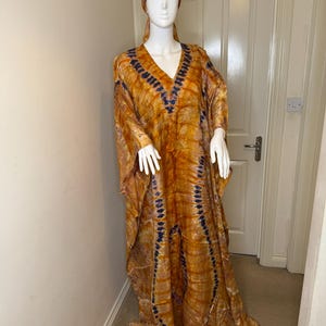 May include: An orange and blue tie-dye kaftan dress with a matching head wrap. The long-sleeved dress has a V-neck and a flowing silhouette. The fabric has a pattern of vertical blue lines against an orange background.