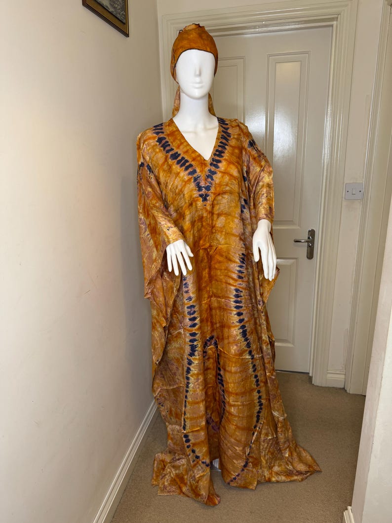May include: An orange and blue tie-dye kaftan dress with a matching head wrap. The long-sleeved dress has a V-neck and a flowing silhouette. The fabric has a pattern of vertical blue lines against an orange background.