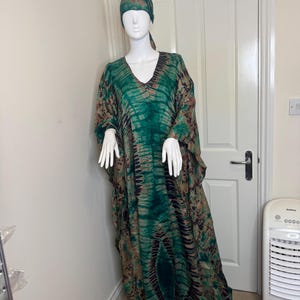 May include: A green and brown tie-dye caftan dress and matching head wrap on a mannequin. The dress has a V-neck and wide sleeves. The fabric has a pattern of dark green and brown vertical stripes.