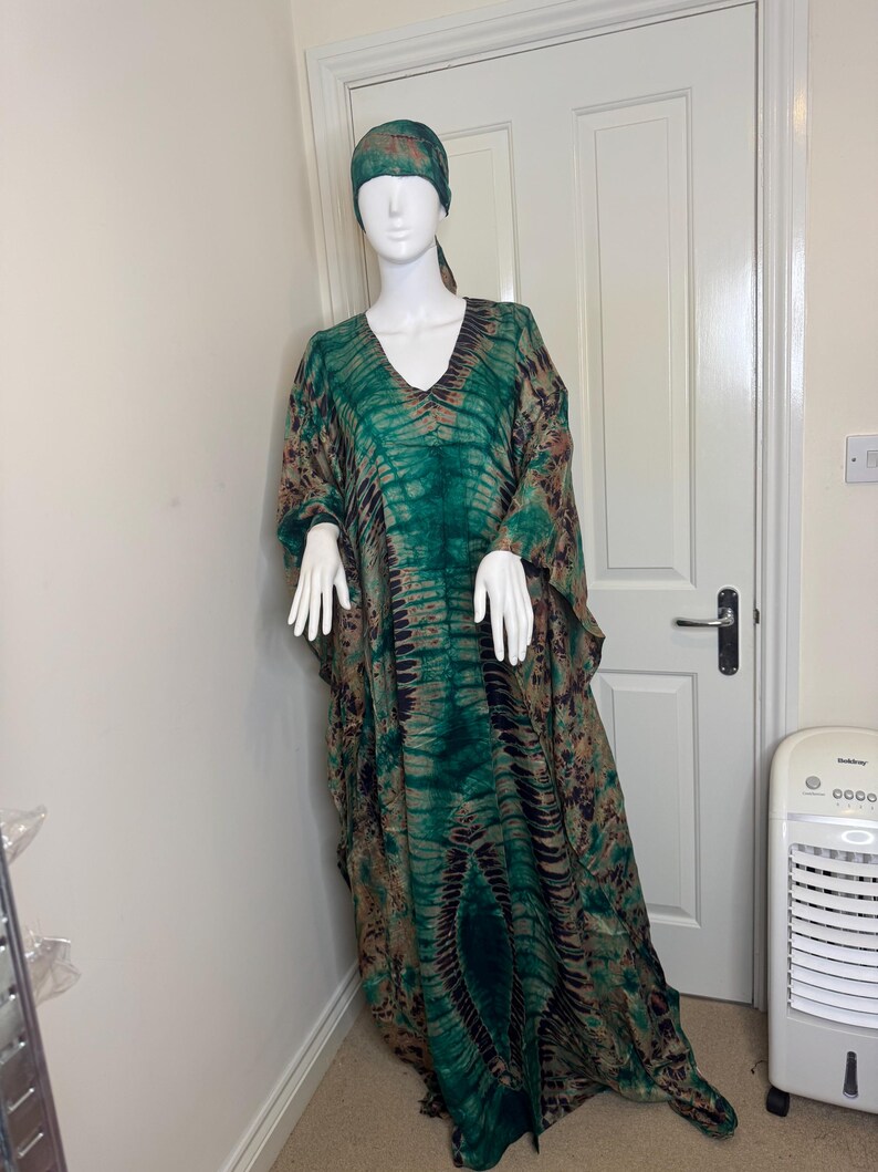 May include: A green and brown tie-dye caftan dress and matching head wrap on a mannequin. The dress has a V-neck and wide sleeves. The fabric has a pattern of dark green and brown vertical stripes.