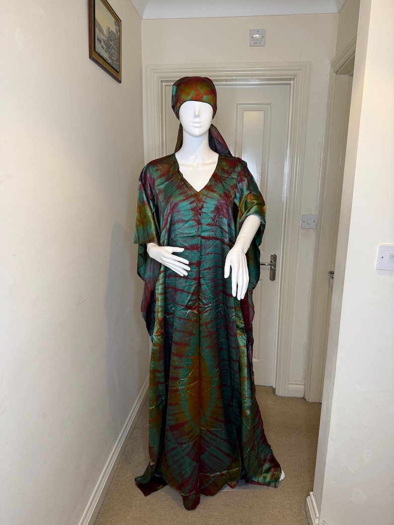 May include: A long, flowing, tie-dye dress and matching headscarf in shades of green, red, and gold. The dress has a V-neck and wide sleeves. The outfit is displayed on a mannequin.