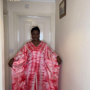 May include: A flowing, tie-dye style caftan in shades of red and white. The V-neck garment has wide sleeves and a loose fit. The caftan is worn by a person in a hallway with a chandelier and a framed picture.