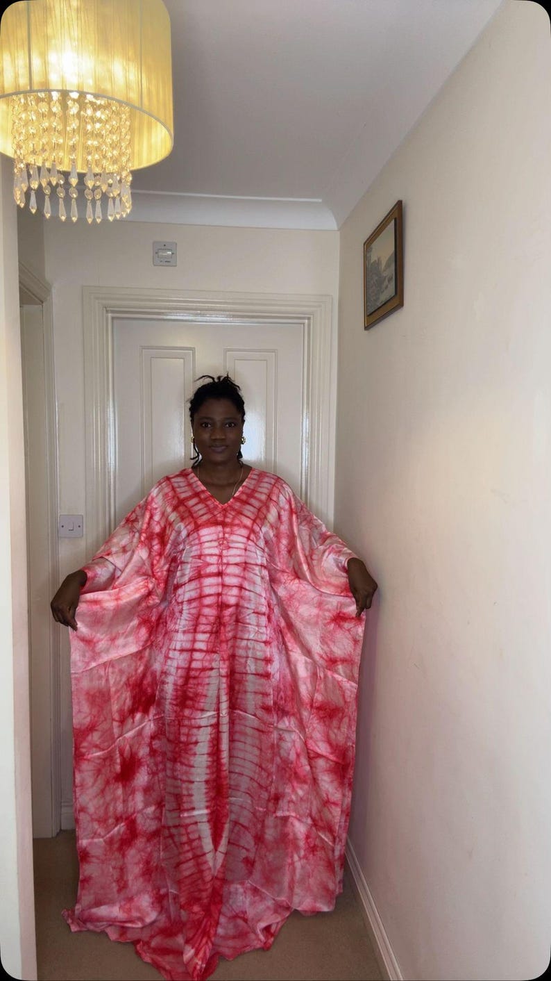 May include: A flowing, tie-dye style caftan in shades of red and white. The V-neck garment has wide sleeves and a loose fit. The caftan is worn by a person in a hallway with a chandelier and a framed picture.