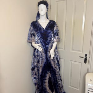 May include: A tie-dye caftan dress and matching head wrap on a mannequin. The dress is a flowing, V-neck design in shades of blue and beige. The fabric has a marbled pattern. The mannequin is positioned in front of a white door.
