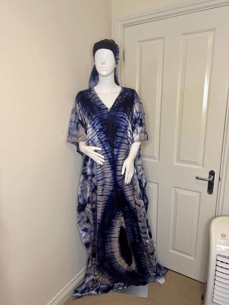May include: A tie-dye caftan dress and matching head wrap on a mannequin. The dress is a flowing, V-neck design in shades of blue and beige. The fabric has a marbled pattern. The mannequin is positioned in front of a white door.