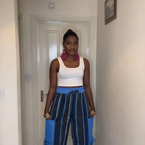 May include: A white tank top paired with wide-leg, high-waisted pants featuring vertical black and blue stripes. The pants have large blue pockets with fringe detailing. Gold bracelets and large pink earrings complete the look.