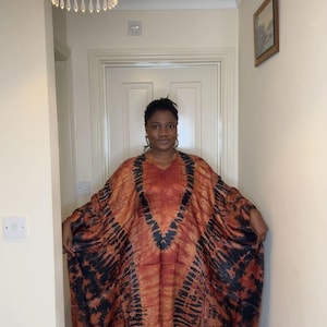 May include: A flowing, V-neck dress in shades of orange and brown with black tie-dye patterns. The dress has wide sleeves and a loose fit. The fabric appears to be lightweight and suitable for warm weather. The dress is worn by a person.
